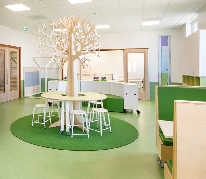 Op de afbeelding zie je een groene ruimte met banken en een tafel om een nepboom.